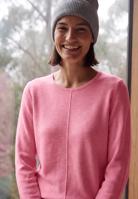 Junge Frau in rosa Pullover und grauer Mütze, die vor einem Fenster lächelt