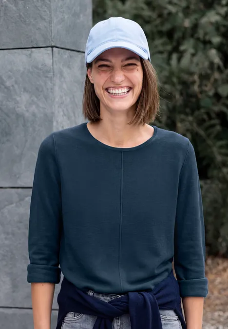 Eine junge Frau mit Baseballcap und blauem Shirt lächelt breit im Freien vor einer Steinwand.