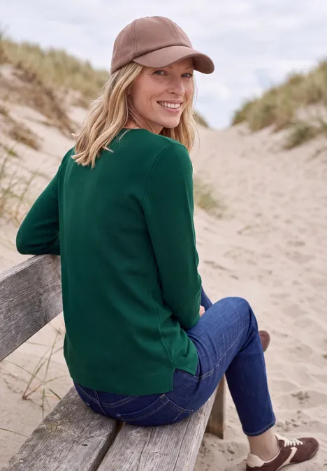 Langarm Pullover mit Rundhals und Struktur dark fir green Frau im grünen Pullover und Jeans sitzt am Strand auf einem Geländer, trägt Cap und lächelt zur Seite.