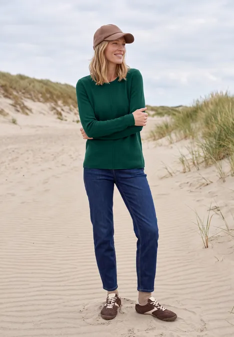 Langarm Pullover mit Rundhals und Struktur dark fir green Frau in grünem Pullover, Jeans und Cap steht entspannt auf einer sandigen Düne mit Grasbüscheln.