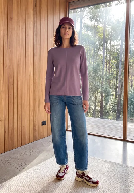 Femme en pull violet, jean bleu, baskets et casquette debout dans un espace lumineux devant de grandes fenêtres.