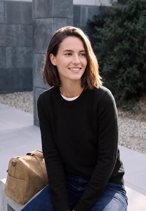 Jeune femme souriante assise dehors avec un sac à dos devant un mur et des arbustes.
