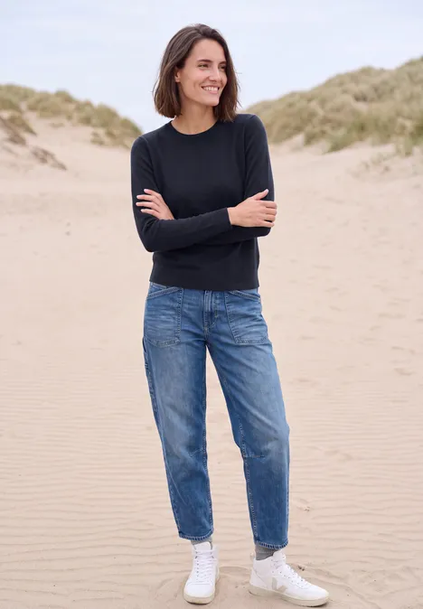 Eine lächelnde Frau mit schwarzem Pullover, blauen Jeans und weißen Sneakers steht an einem Sandstrand vor Dünen.
