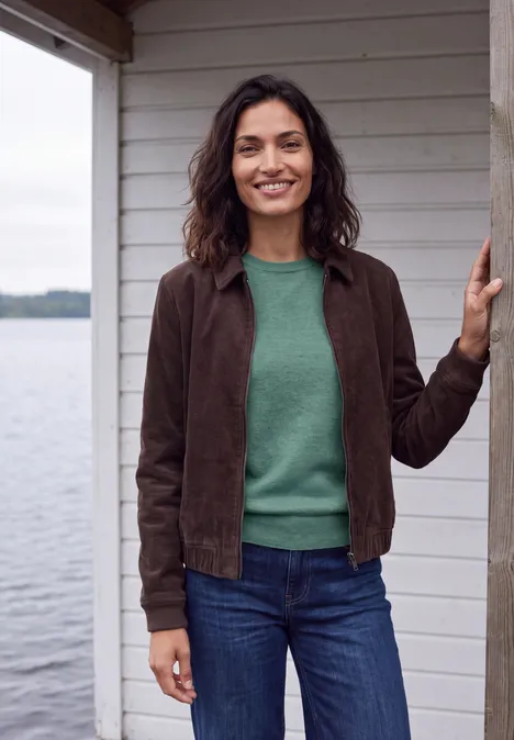 Lächelnde Frau in grünem Pullover, brauner Jacke und Jeans steht an einem Holzhaus am Wasser.