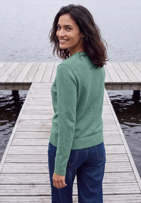 Eine Frau in grünem Pullover und Jeans steht auf einem Steg am Wasser und lächelt über die Schulter.
