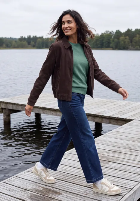 Frau in grünem Pullover und brauner Jacke spaziert lachend auf einem Steg am See.