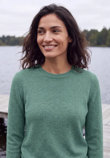Eine lächelnde Frau im grünen Pullover vor einer See-Kulisse.