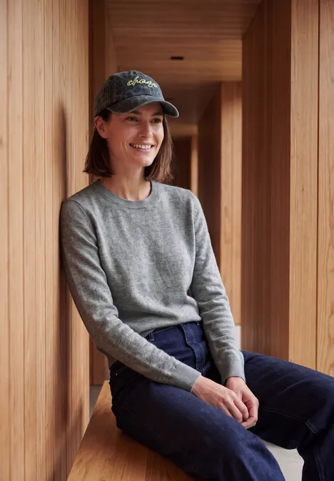 Eine Frau sitzt lächelnd in einer Holzstruktur und trägt grauen Pullover, blaue Hose sowie Baseballkappe.