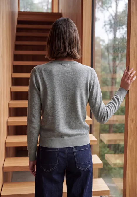 Eine Person in grauem Pullover und Jeans steht mit dem Rücken zur Kamera vor einer Holztreppe.