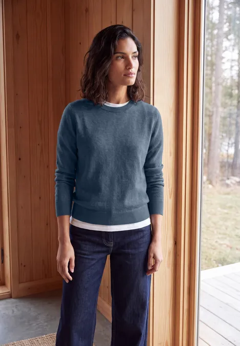 Femme en pull bleu et jean debout dans une pièce en bois clair, regard tourné vers l’extérieur.