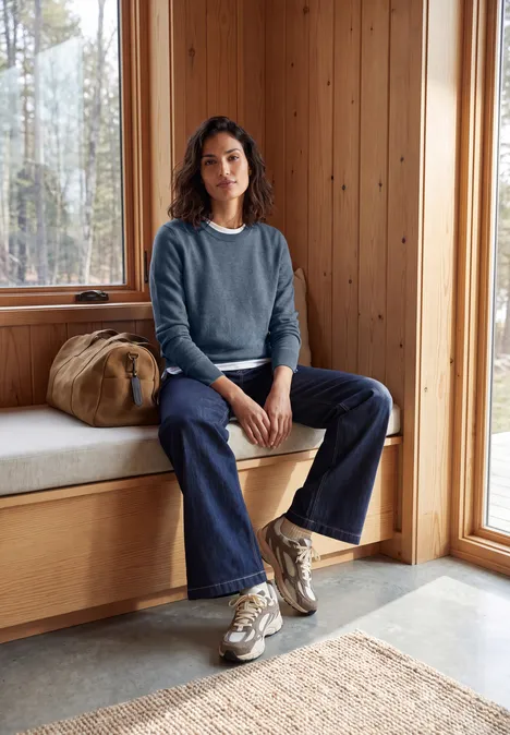 Une femme en pull, jean et baskets est assise tranquillement sur un banc en bois près d’une grande fenêtre.