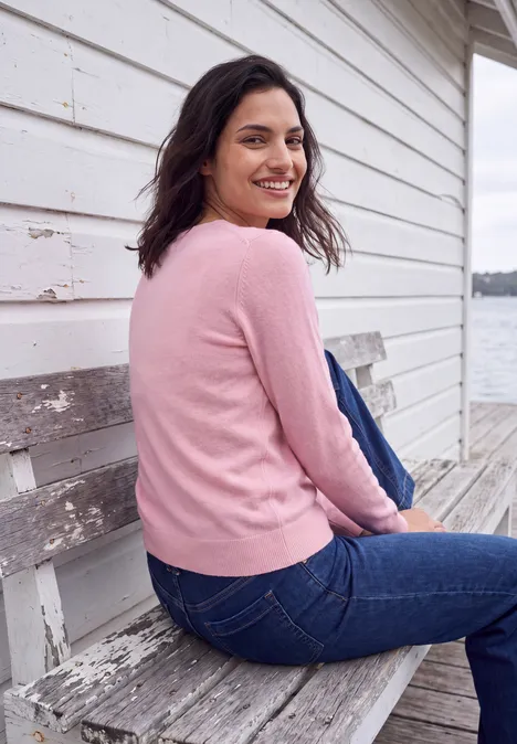 Frau in rosa Pullover und Jeans sitzt auf hölzerner Bank am Wasser und lächelt zur Kamera.