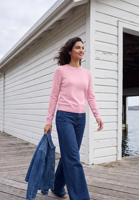 Frau in rosa Pullover und Jeans spaziert mit Jeansjacke an einem Steg vor Bootshaus.
