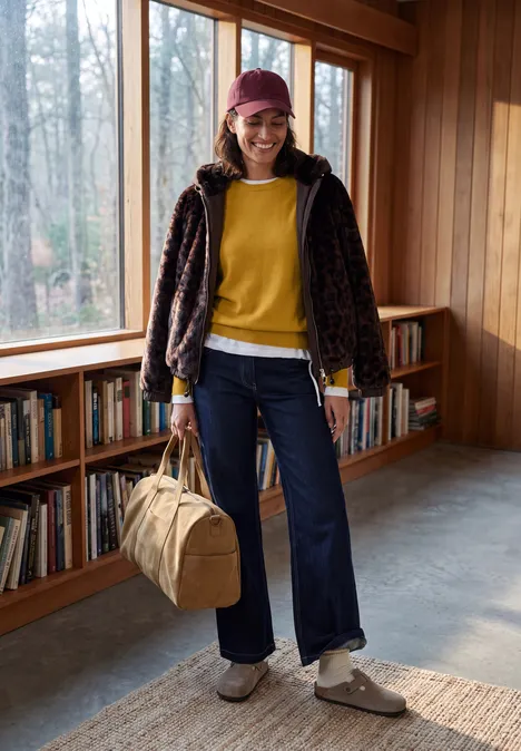 Trui met ronde hals golden honey melange Person in gelbem Pullover, Jeans, gemusterter Jacke und Beanie hält Reisetasche in Holzraum mit Bücherregal