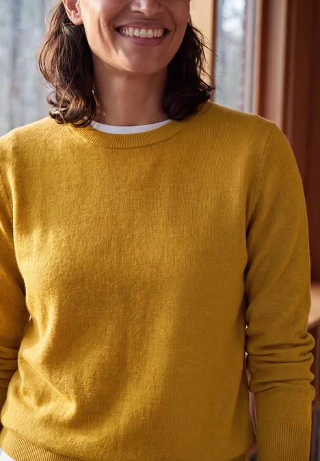 Trui met ronde hals golden honey melange Eine lächelnde Frau trägt einen gelben Pullover in einem gemütlichen Innenraum.