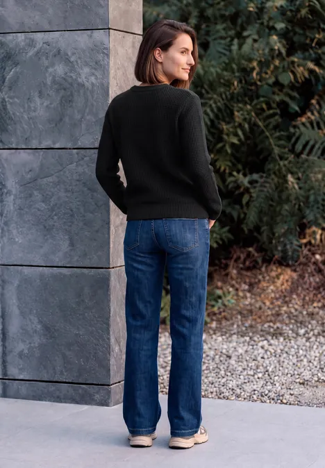 Femme en pull noir et jean bleu, appuyée contre un mur gris, regarde un sol caillouteux et des buissons.