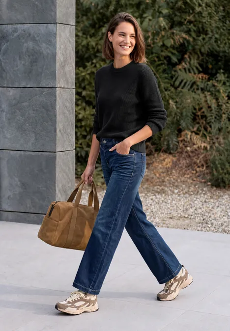 Une femme souriante en pull noir, jean bleu et baskets marche en plein air avec un sac marron.