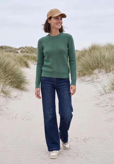 Frau in grünem Pullover und Jeans spaziert am Strand zwischen Dünen