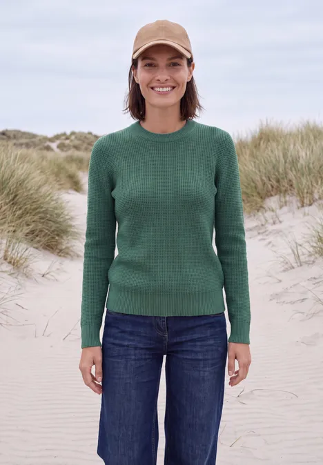 Eine Frau trägt grünen Pullover, Jeans und beige Mütze, lächelt vor Dünen am Strand.