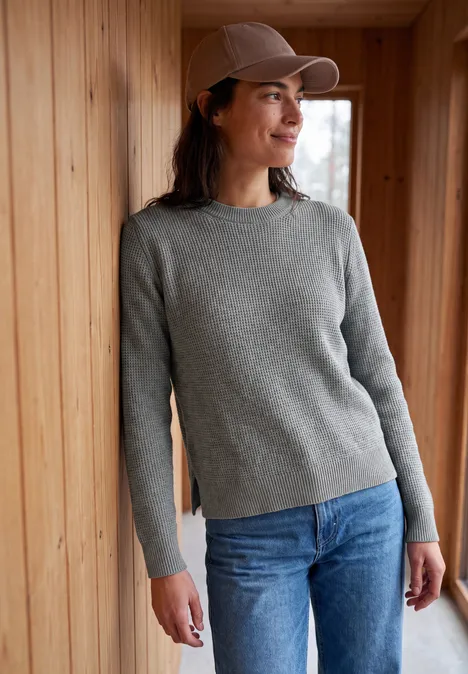 Eine Frau lehnt in grauem Pullover, blauer Jeans und Cap an einer Holzverkleidung und lächelt.