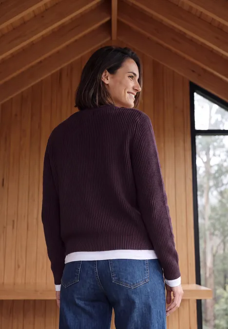 Frau mit lila Pullover und Jeans blickt über die Schulter in einem Holzhaus.