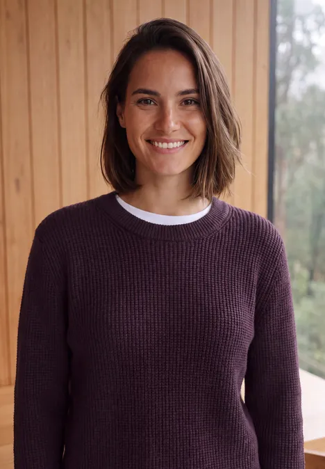 Lächelnde Frau mit kurzen dunklen Haaren in violettem Pullover vor Holzverkleidung.