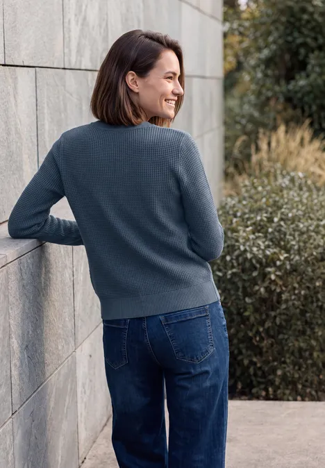 Trui met ronde hals cotton blue melange Eine lächelnde Frau in grauem Pullover und blauer Jeans lehnt sich an eine Steinwand und schaut zur Seite.