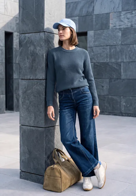 Trui met ronde hals cotton blue melange Frau lehnt an grauer Wand, trägt dunklen Pulli, weite Jeans, weiße Sneaker und Cap, neben ihr liegt eine Reisetasche.