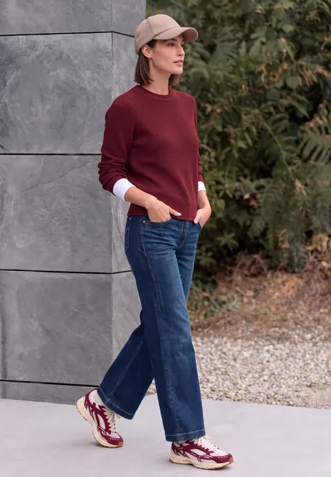 Trui met ronde hals rhubarb melange Person trägt bordeauxfarbenen Pullover, weite Jeans, Cap und Sneakers, steht vor einer Steinwand und Herbstlaub.