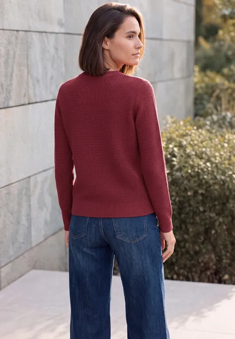 Trui met ronde hals rhubarb melange Frau im roten Pullover und Jeans steht vor einer Steinmauer und Büschen im Freien.