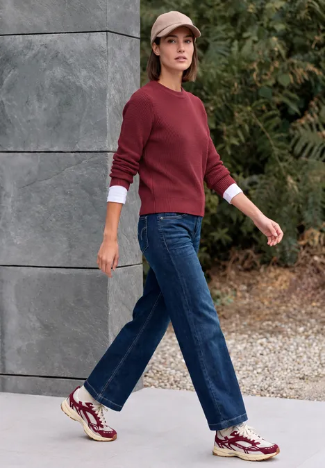 Trui met ronde hals rhubarb melange Frau in rotem Pullover, Jeans, Baseballcap und Sneakern geht draußen an grauer Wand entlang.