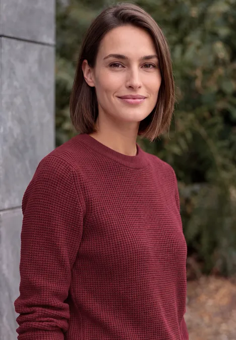 Trui met ronde hals rhubarb melange Eine Frau mit kinnlangen braunen Haaren und bordeauxrotem Pullover im Freien.