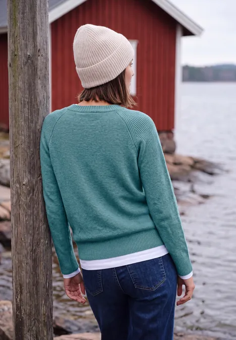 Person mit grüner Strickjacke und Beanie steht rückwärts am Wasser vor einem roten Gebäude.