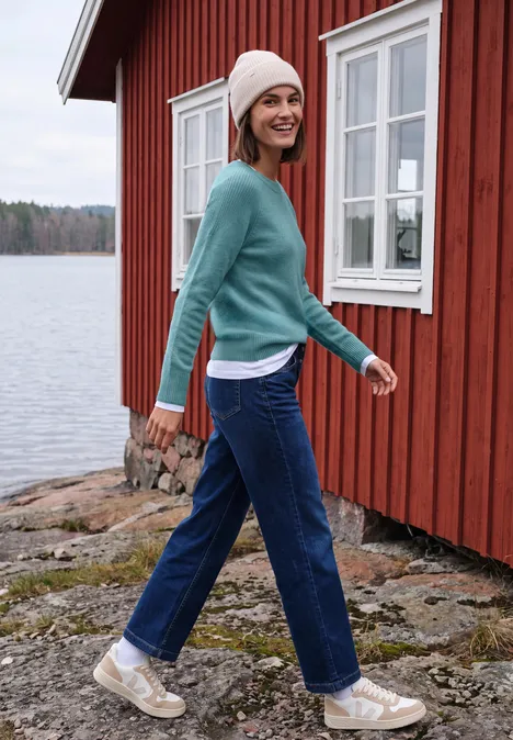 Eine Frau in Jeans und Pullover spaziert lächelnd am Seeufer vor einem roten Haus.