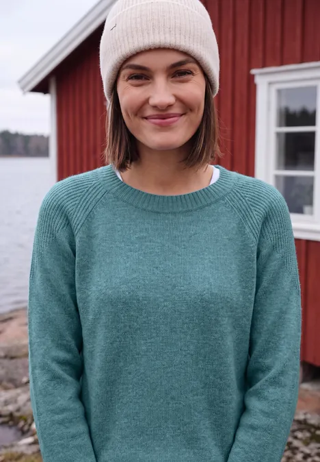 Eine lächelnde Frau mit Strickmütze und Pullover vor einem roten Haus am Wasser.