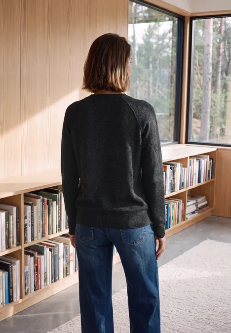 Pull col rond à textures contrastées black melange Person steht mit dem Rücken zur Kamera in einem Holzraum mit Bücherregalen und großen Fenstern.