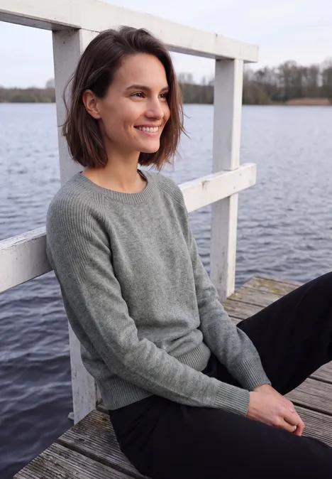 Een glimlachende vrouw in een grijze trui zit ontspannen op een steiger aan het meer.