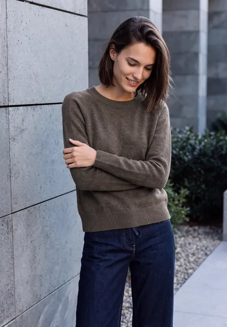 Jeune femme aux cheveux bruns, vêtue d’un pull et d’un jean, appuyée et souriante devant un mur.