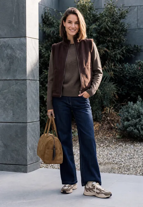 Femme en veste marron, jean bleu foncé et baskets, tenant un sac camel devant un bâtiment moderne.