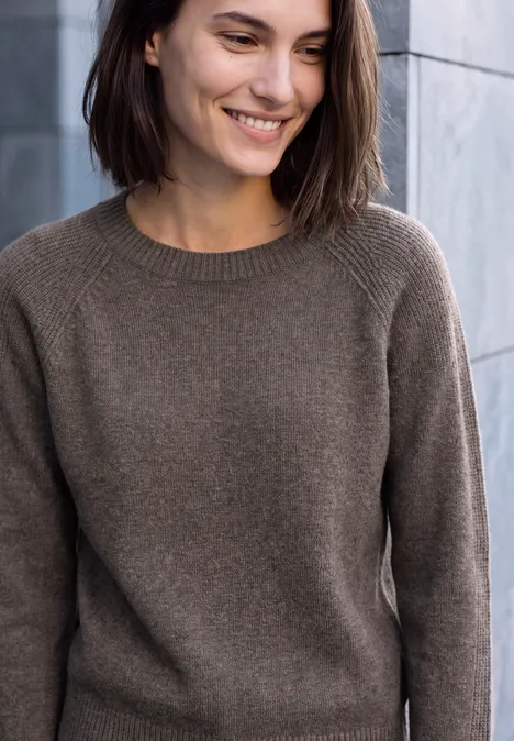 Une femme souriante en pull marron devant un mur de pierre.