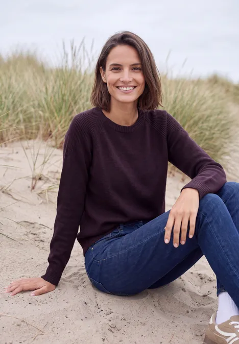 Pullover mit Rundhals und Strukturmix eggplant red melange Eine lächelnde Frau sitzt entspannt im Sand vor Dünengras, trägt einen dunklen Pullover und blaue Jeans.