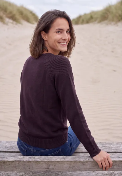 Pullover mit Rundhals und Strukturmix eggplant red melange Eine Frau sitzt auf einer Holzbank am Strand, schaut lächelnd über die Schulter zu den Dünen.