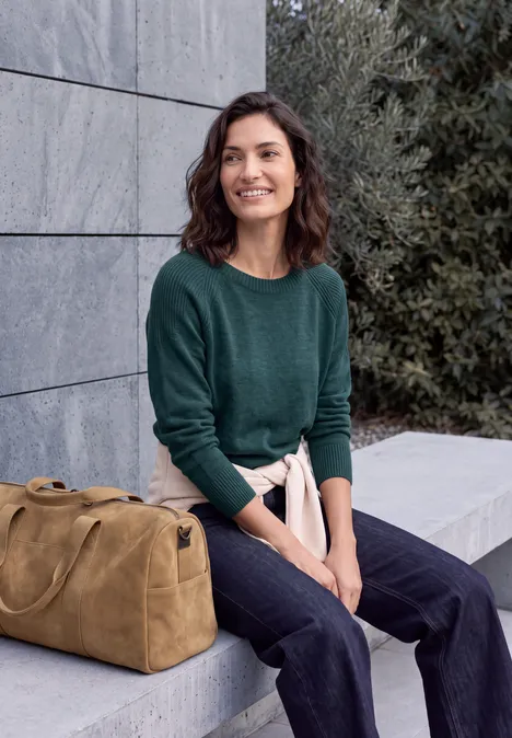 Trui met ronde hals en structuurmix dark sea green melange Junge Frau in grünem Pullover und Jeans sitzt vor einer Mauer, lächelnd neben einer braunen Reisetasche.