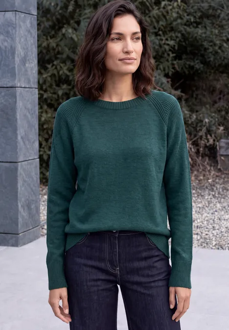 Trui met ronde hals en structuurmix dark sea green melange Frau in grünem Pullover und Jeans steht vor Mauer und Büschen im Freien.