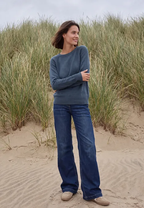 Pullover mit Rundhals und Strukturmix cotton blue melange Frau steht am Strand vor Dünen, trägt blauen Pullover und Jeans, lächelnd im Wind.