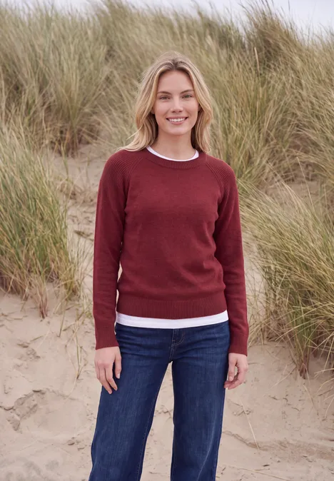 Eine lächelnde Frau in rotem Pullover und Jeans steht am Strand vor hohen Dünengräsern.