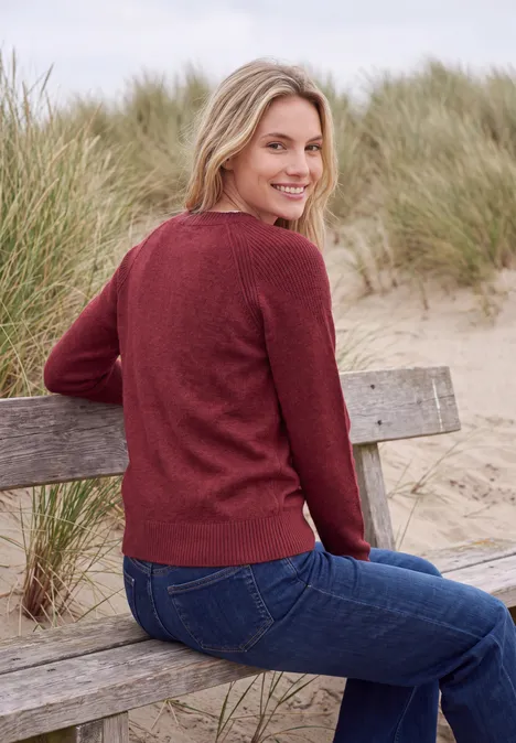 Frau mit rotem Pullover und Jeans sitzt auf einer Holzbank in den Dünen und lächelt.