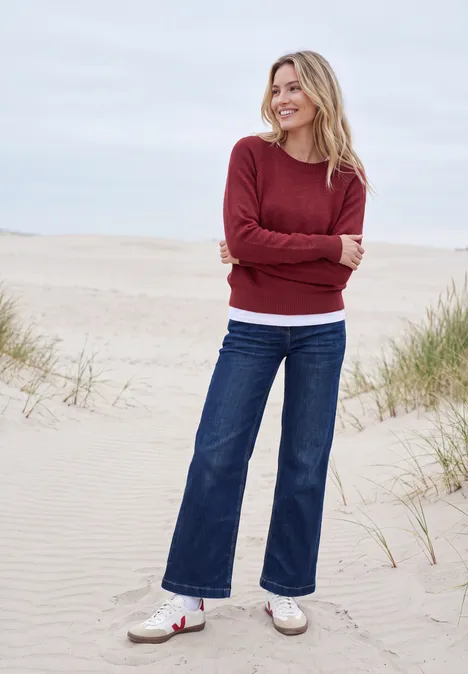 Frau lächelt in rotem Pullover auf einer Dünenlandschaft am Strand.