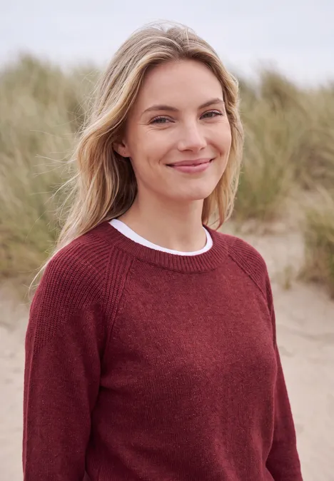 Eine lächelnde Frau in rotem Pullover steht vor grasbedeckten Dünen am Meer.