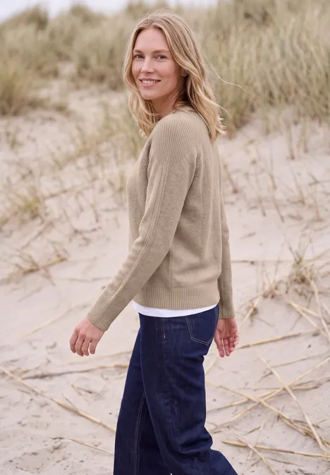 Eine lächelnde Frau in beigem Pullover und dunklen Jeans spaziert am Strand durch Dünen.